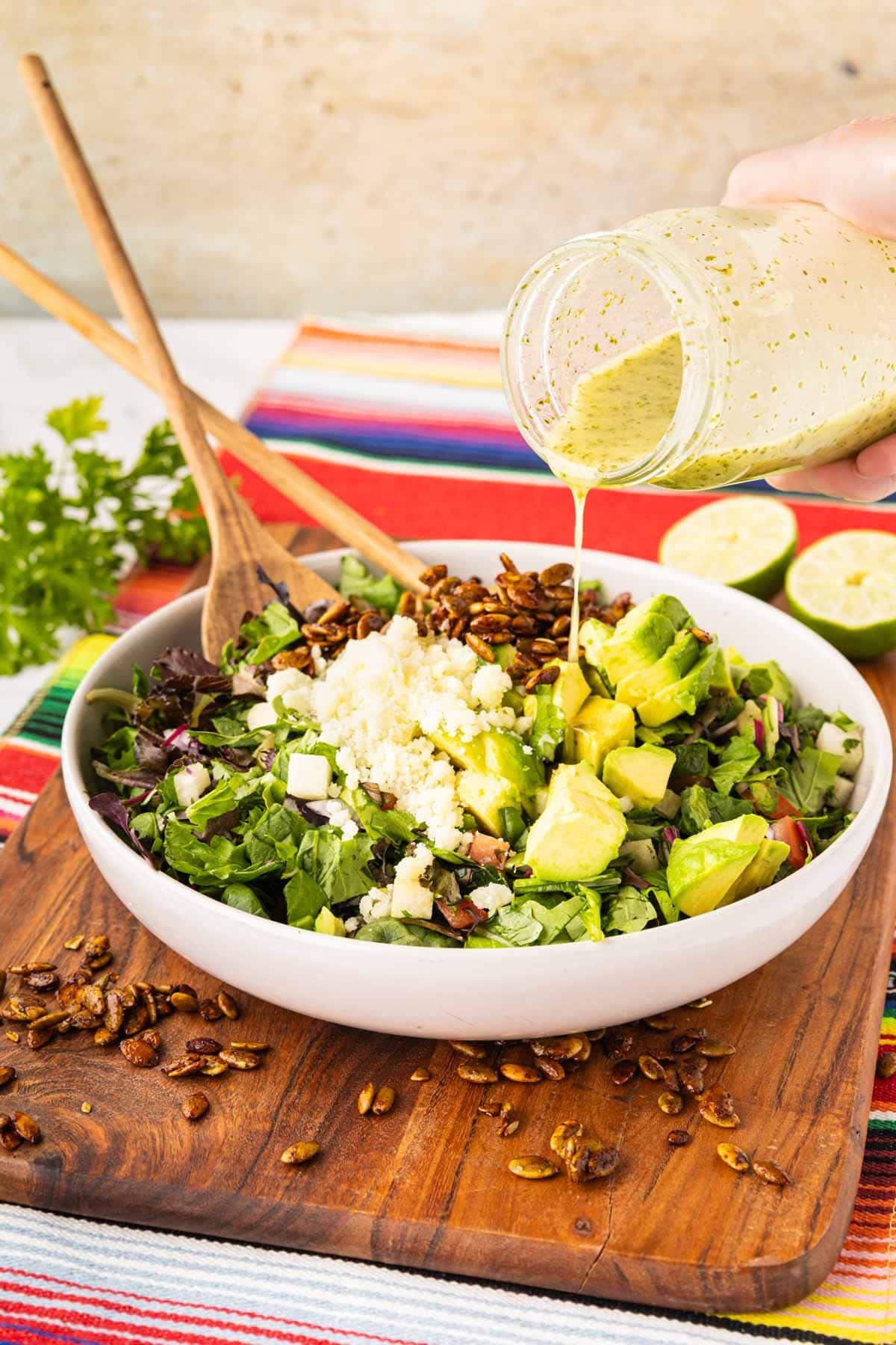 Pouring cilantro, lime vinaigrette on the Mexican, chopped salad, topped with cheese, avocado, and spiced pepitos.