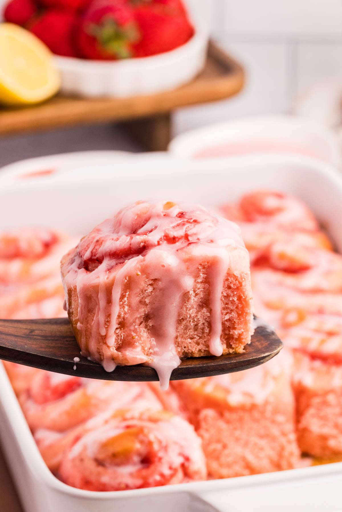 Strawberry cinnamon roll on a wooden spatula hovering over the rest of the rolls.