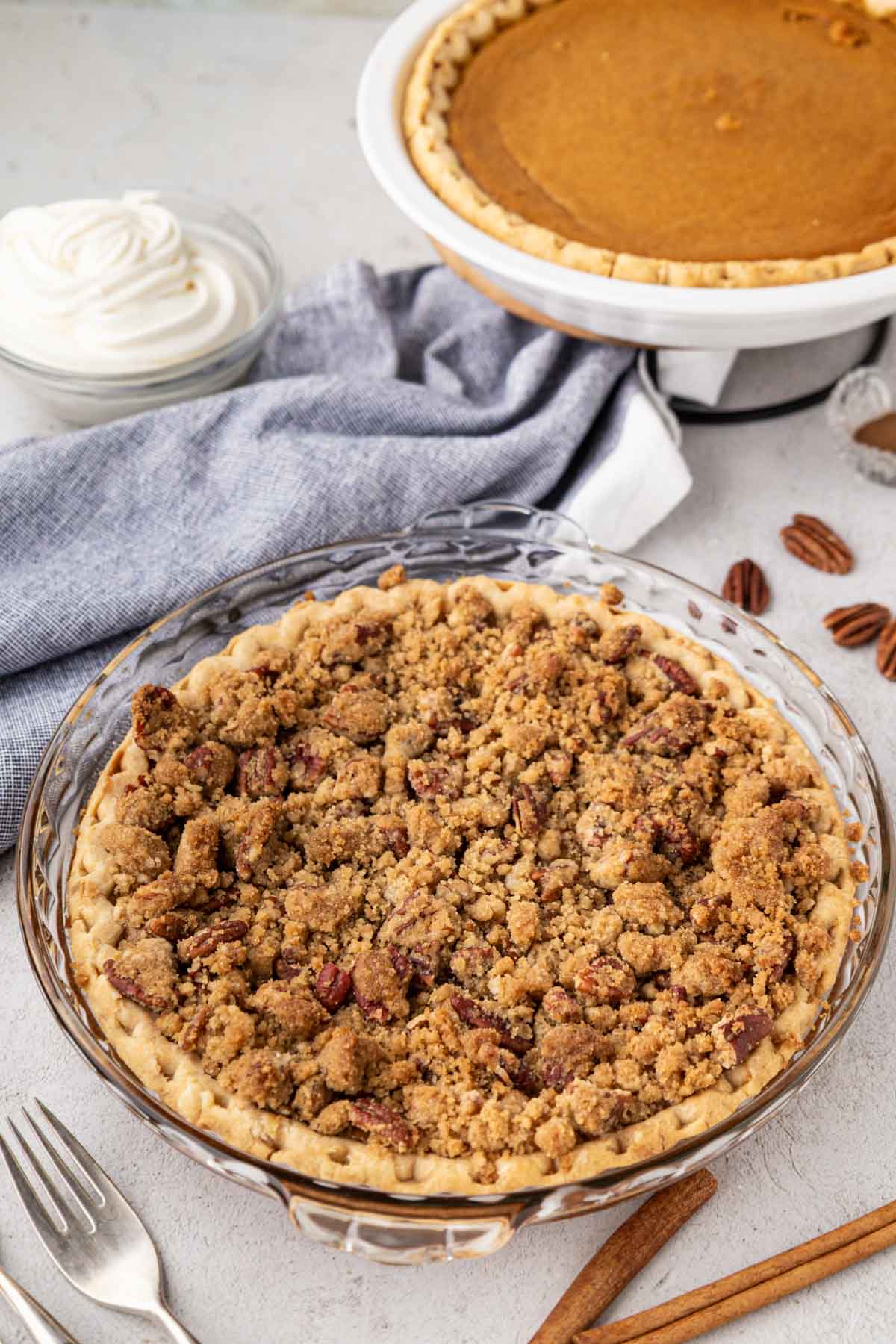 A whole sweet potato pie with a pecan crumb topping on a table with a sweet potato pie in the background without a topping and whipped cream.