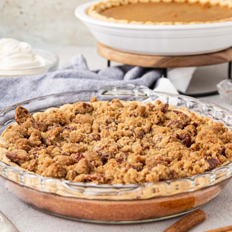 A sweet potato pie with a pecan crumb topping on a table with a sweet potato pie in the background without a topping and whipped cream.