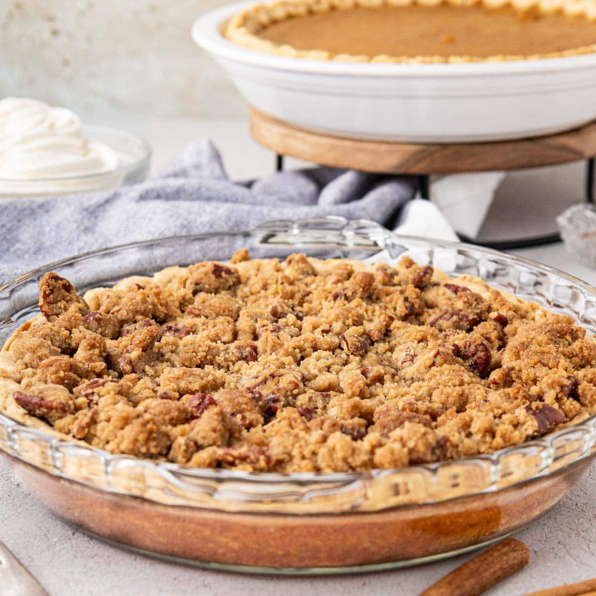 A sweet potato pie with a pecan crumb topping on a table with a sweet potato pie in the background without a topping and whipped cream.