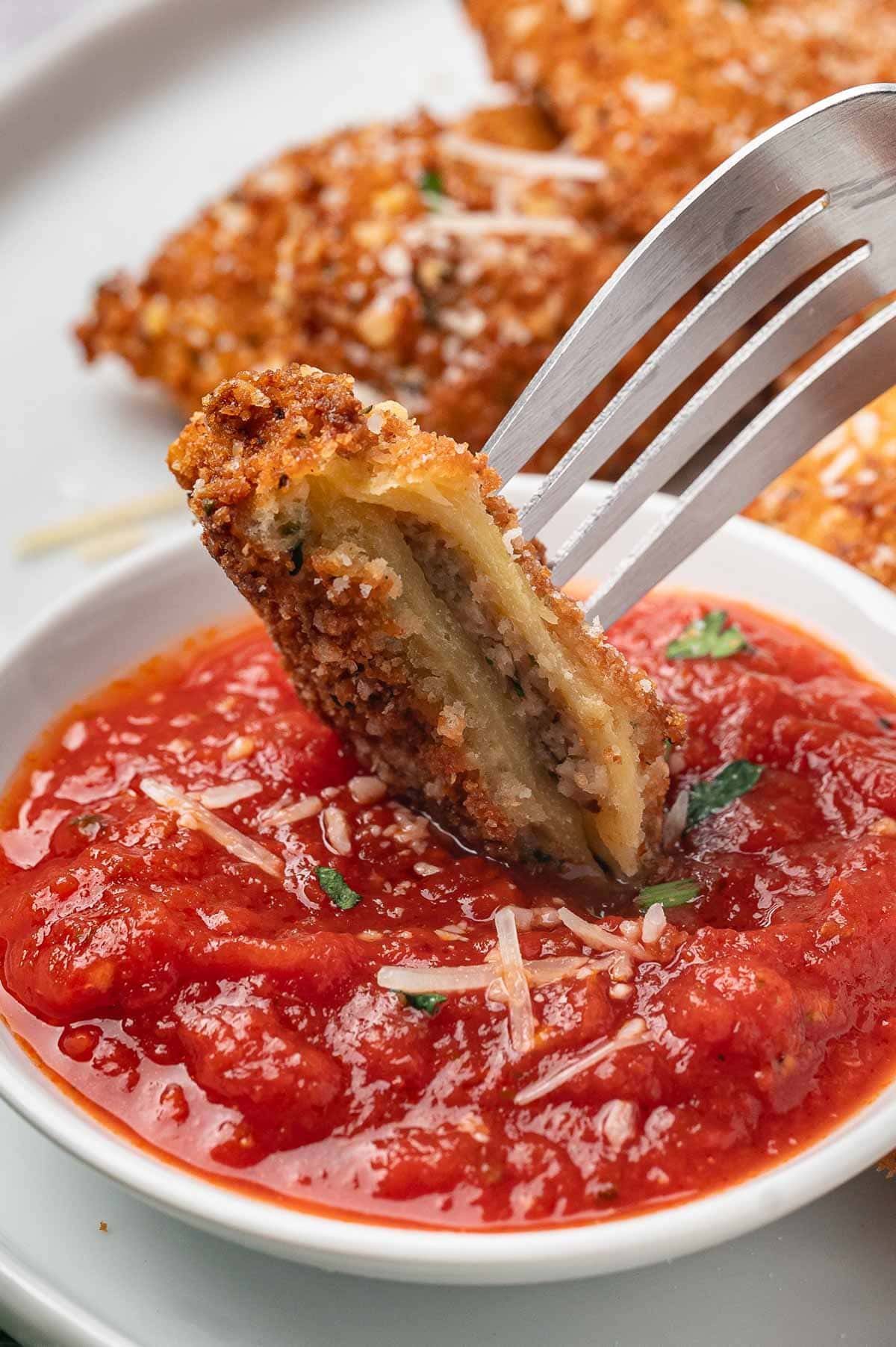 Fork with a bite of toasted ravioli on it being dipped into tomato sauce.