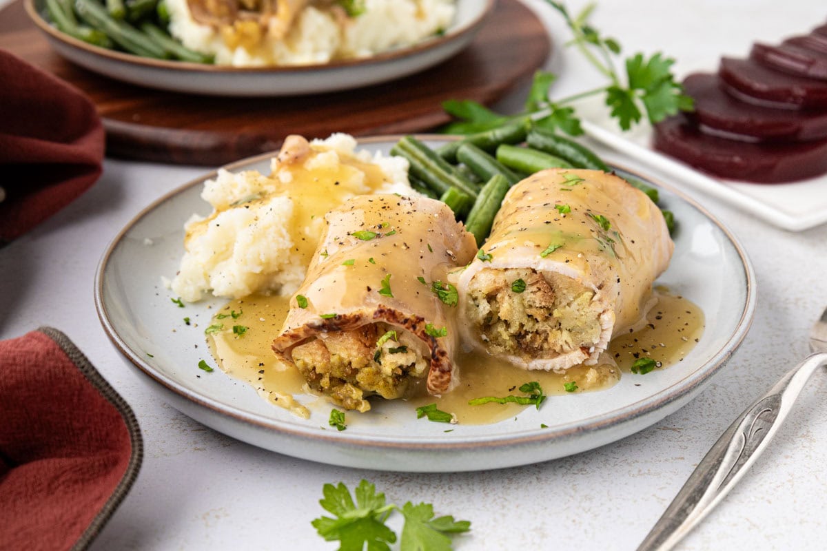 A turkey and stuffing roll up sliced in half with gravy spoon over it served with a side of mashed potatoes and green beans.