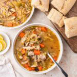 Overhead shot of chicken cabbage soup in a white bowl with bread on a wooden board.