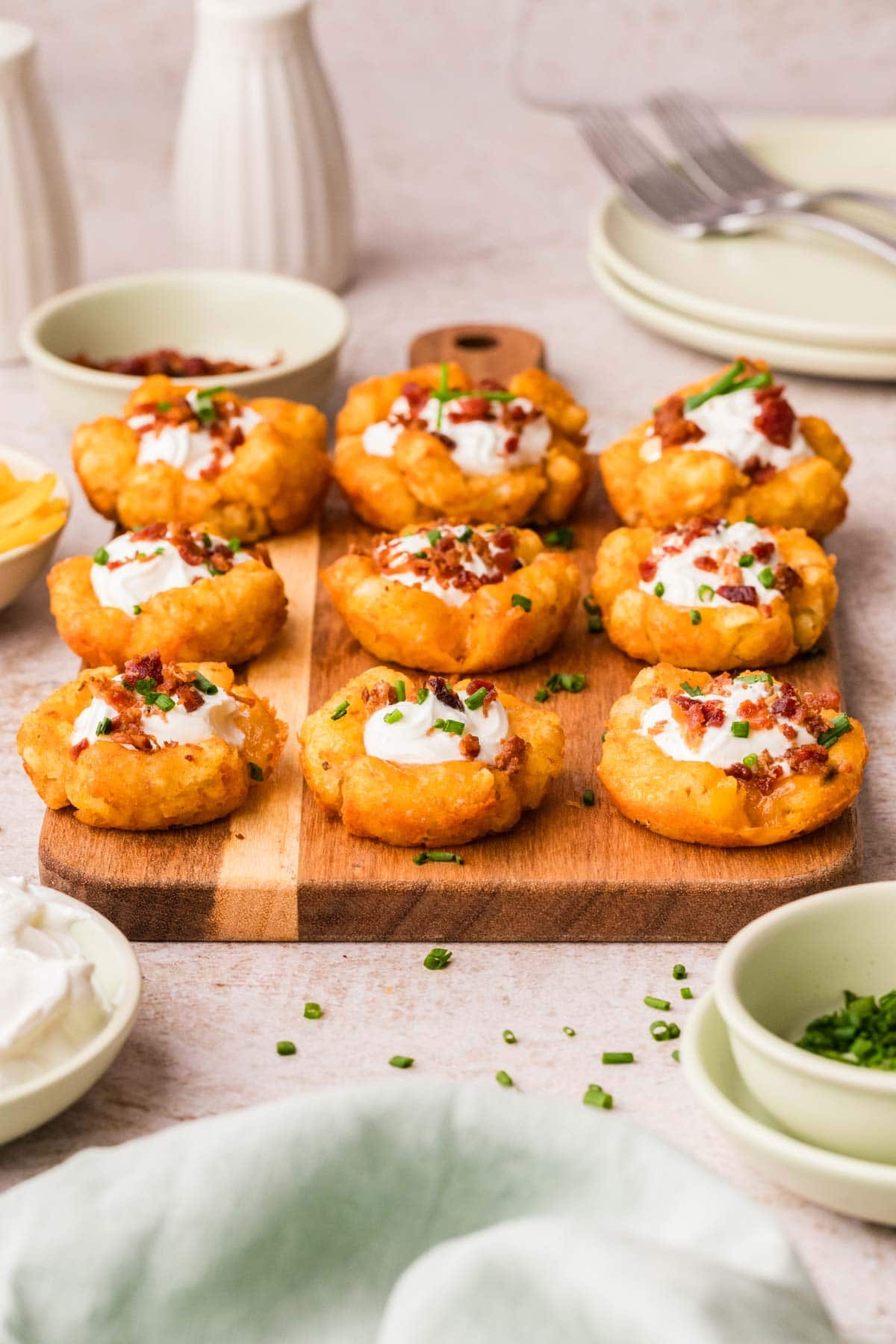 Tater tot cups lined up on a wooden board, all garnished with bacon and chives.