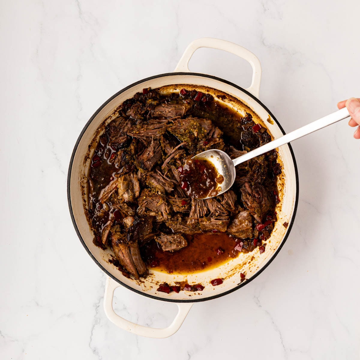 Hand spooning the juices of the pot roast onto the shredded meat.