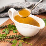 Close up of a metal ladle scooping up pork gravy from a white gravy boat.