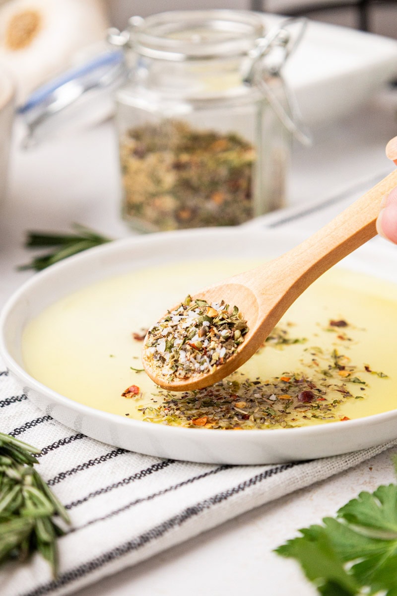  white appetizer plate with a spoon adding Italian Herbs and mixing with olive oil for a dip.