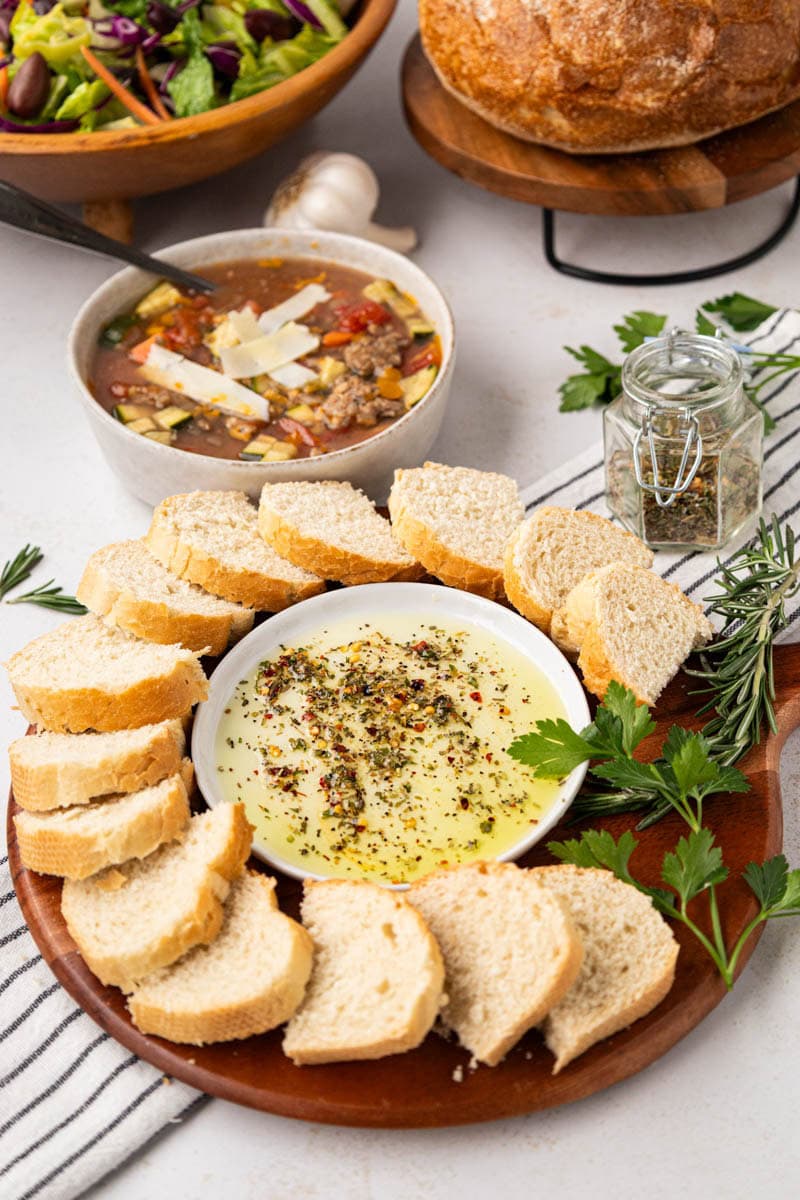 A wooden board with sliced of Italian bread circled around a small plate with herbed dipping oil.