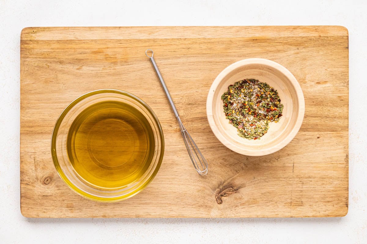 Herbs mixed in a bowl with tiny whisk and a bowl of olive oil.