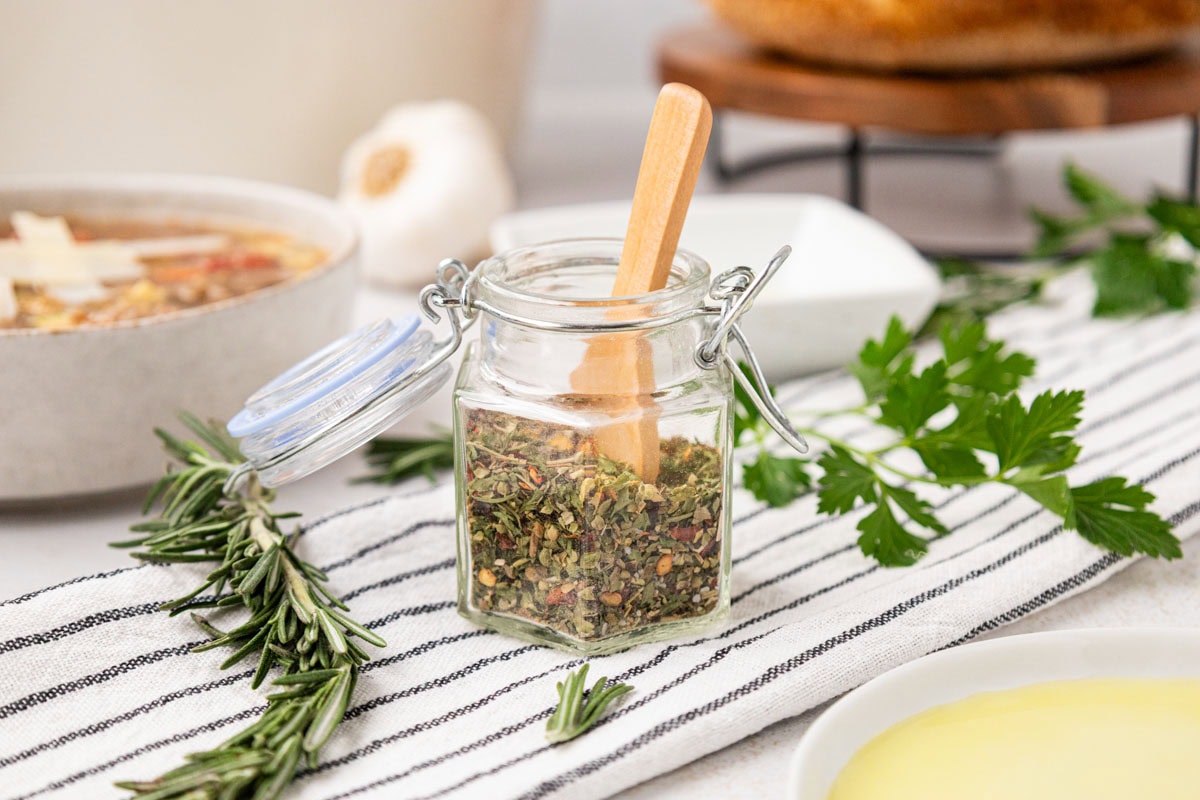 Italian Herb blend in a jar with a wooden spoon. set on a tablescape with fresh herbs.