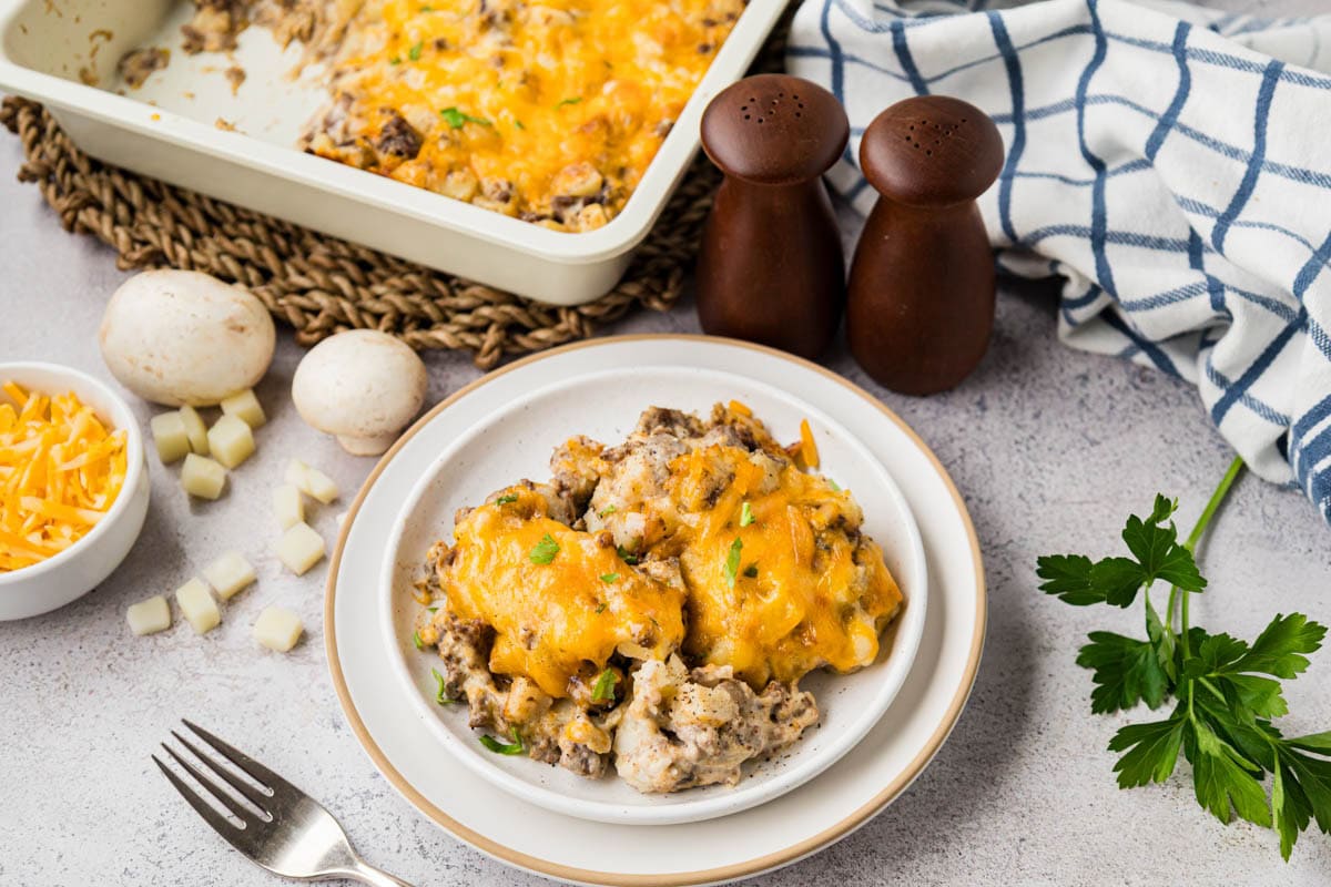Serving of hamburger hash brown casserole on a plate with a baking dish in the background.