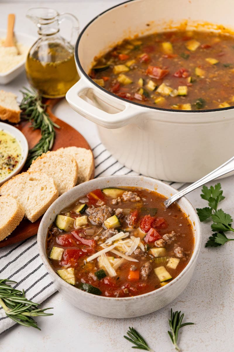 A white bowl filled with sausage and lentil soup with bread and dipping oil along with the soup pot in the background.