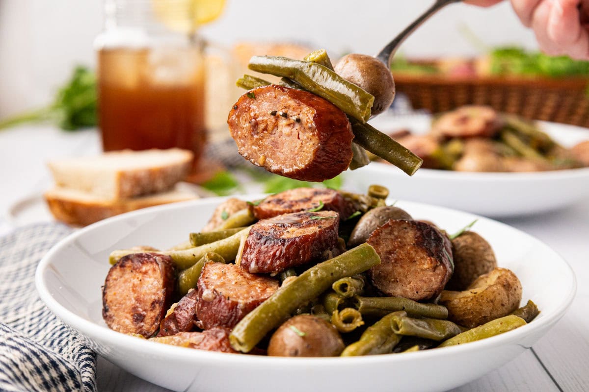 Hand holding a fork with a bite of the crockpot sausage, potatoes and green beans on it.