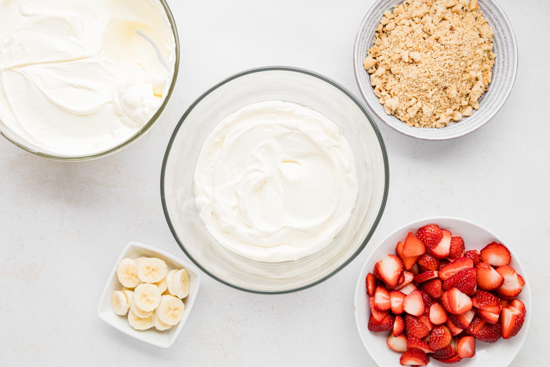 A layer of pudding mixture in the bottom of a trifle dish. With sliced strawberries, slice bananas, crushed cookies and whipped topping  in seperate bowls ready to make layers.