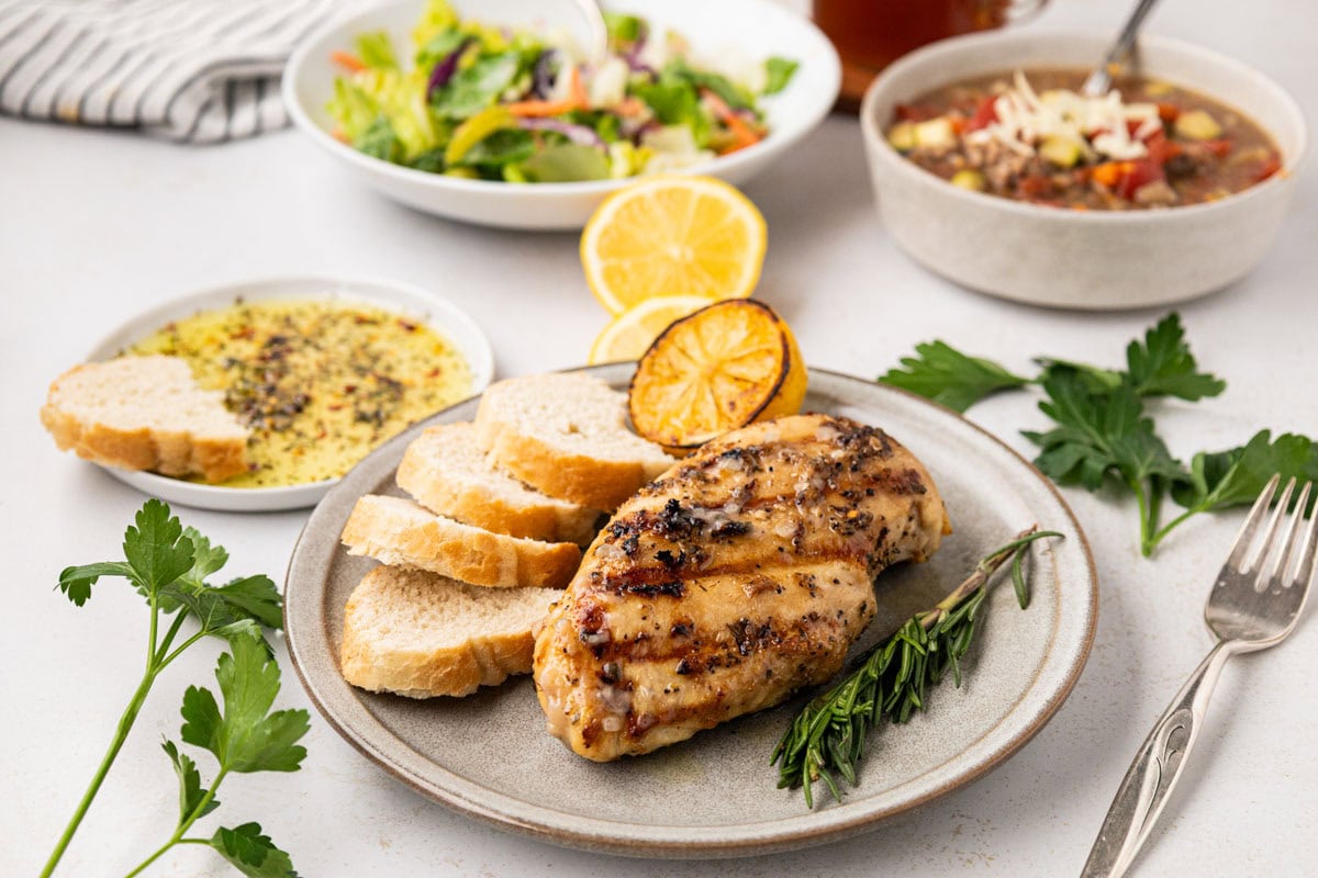 Italian grilled chicken on a plate with bread slices a charred lemon and herbs with soup and salad in the background.