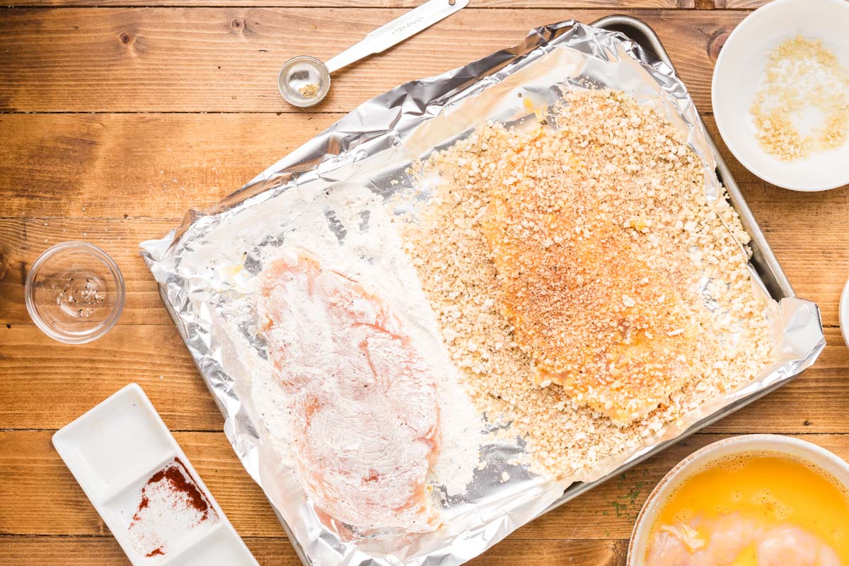 Chicken cutlets being dredged and breaded on a sheet pan.