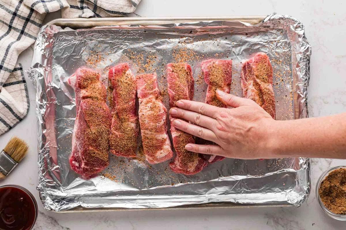 Patting rib rub into ribs on a lined baking sheet.