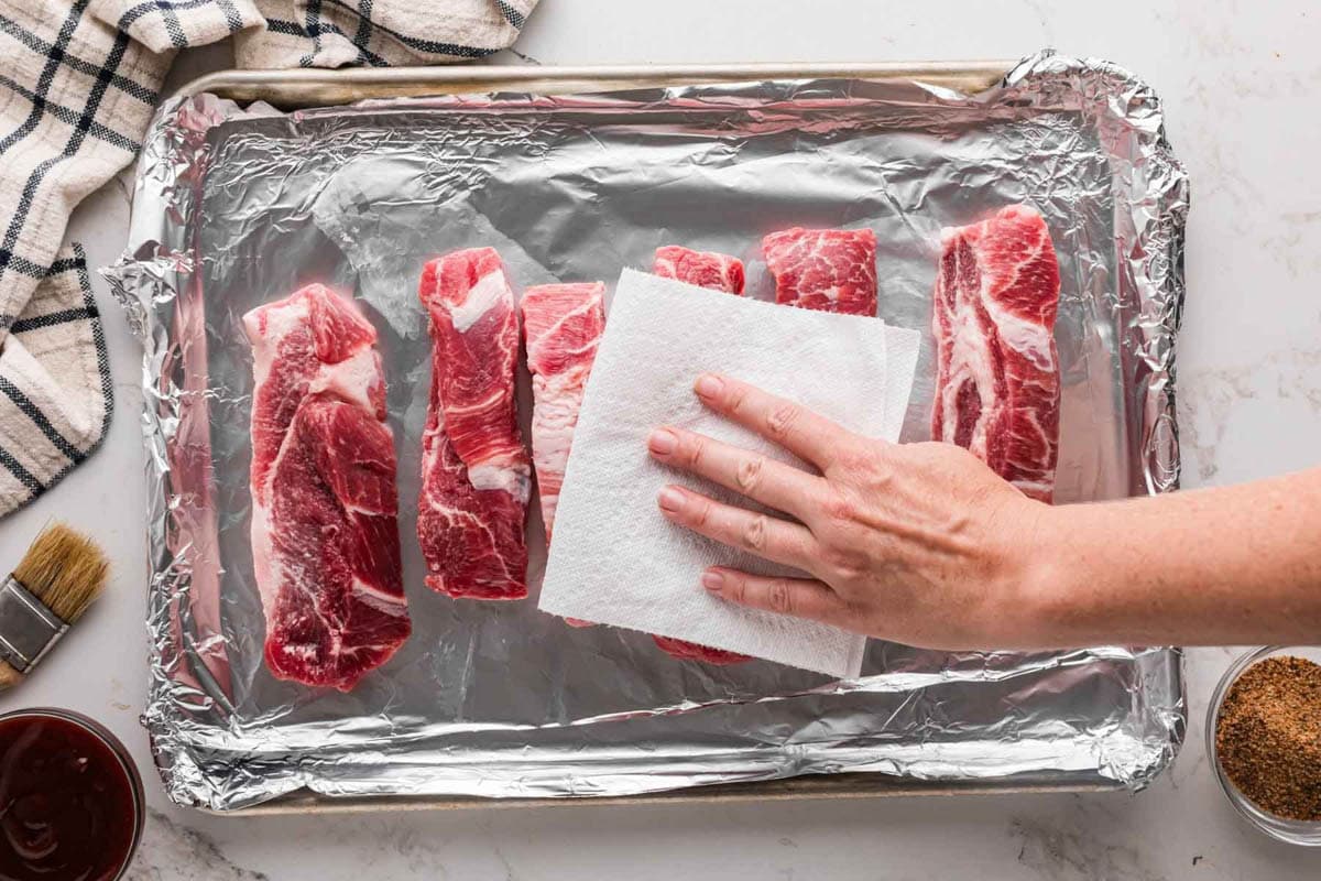 Patting pork ribs dry with a paper towel on a lined baking sheet.
