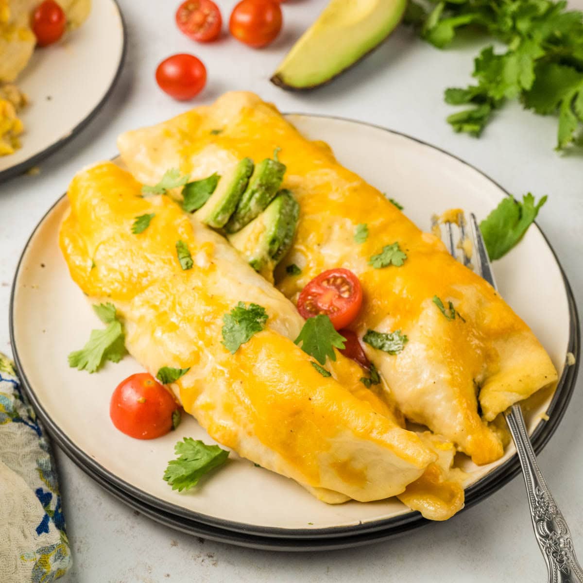 Two breakfast enchiladas on a plate garnished with cilantro diced avocado, and tomatoes.