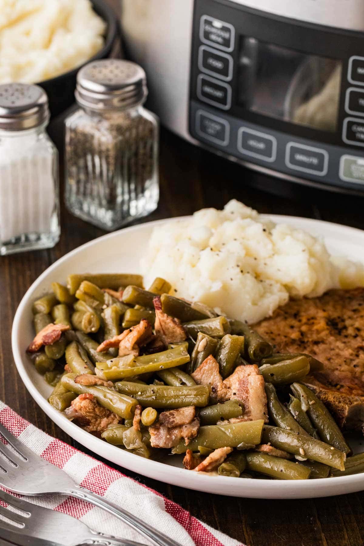 A white plate with slow cooker green beans and bacon served with mashed potatoes and fried pork chops with a slow cooker in the background.