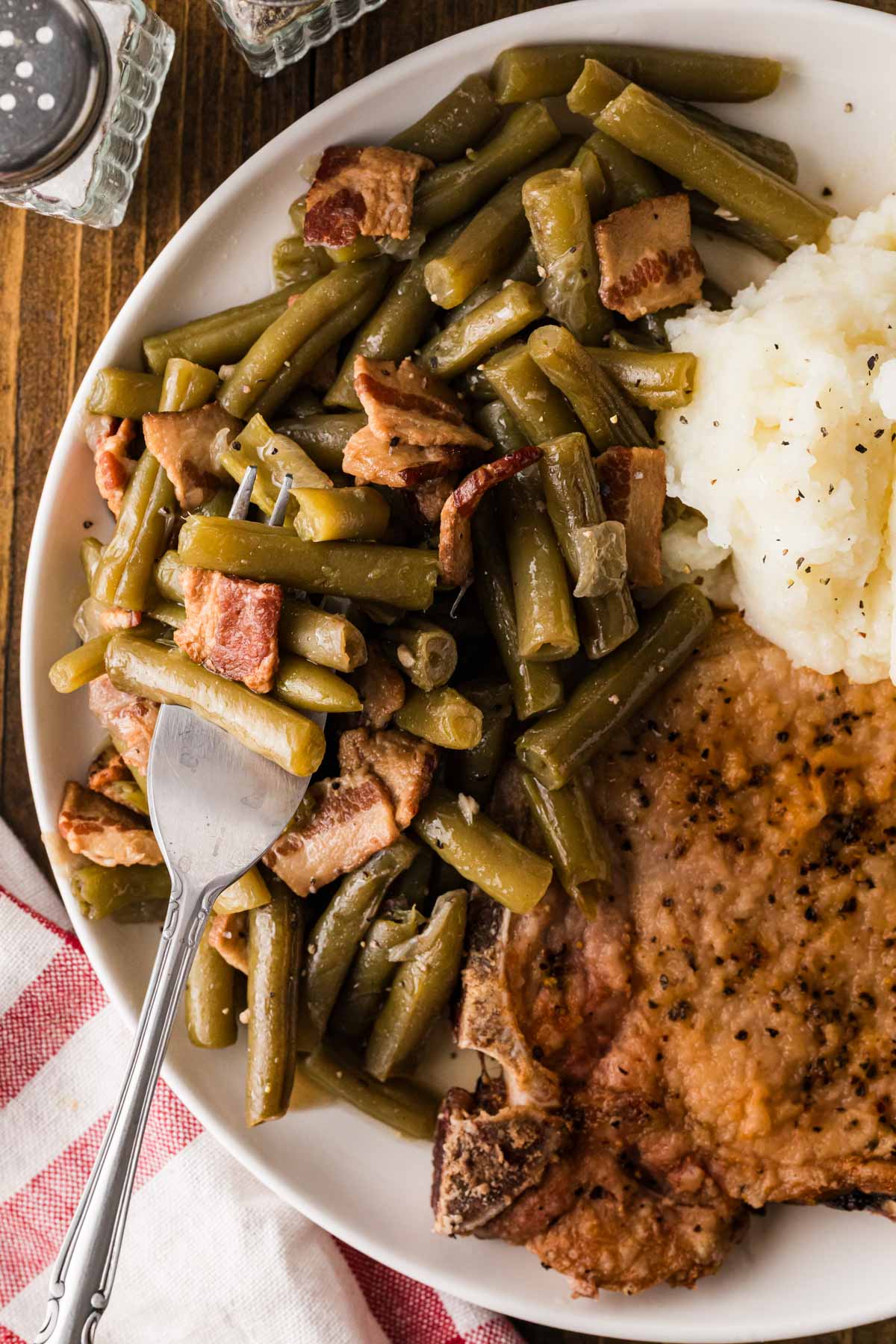 An overhead close up shot of a fork getting a bite of the green beans off of a plate with mashed potatoes and pork chops.