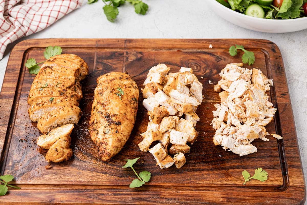 Four chicken breast on a wooden cutting board; one slice, one whole, one chopped and one shredded.