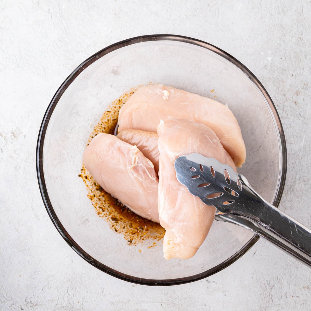 A pair of tongs adding chicken breast to marinade in a bowl.