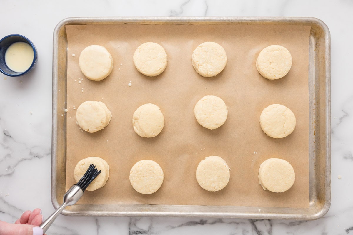 English scones being brushed with egg wash on a baking sheet lined with parchment paper.
