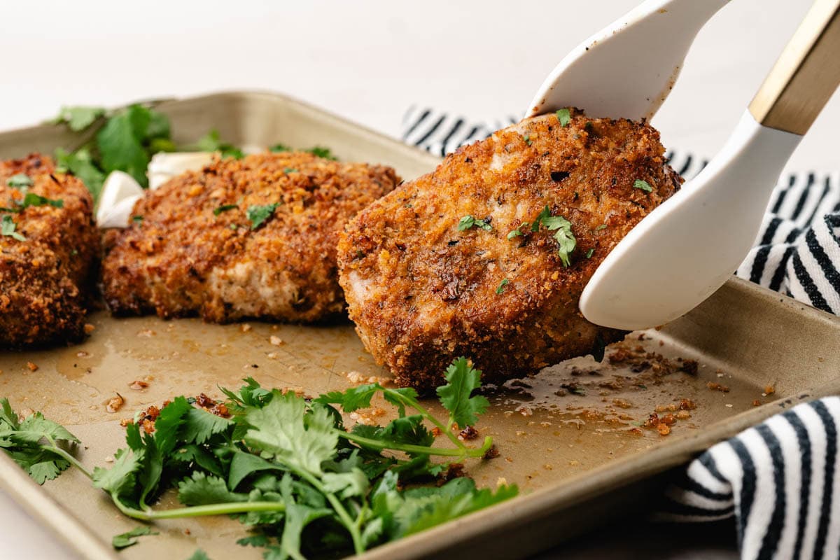 Tongs grabbing a parmesan crusted pork chop from the baking sheet.
