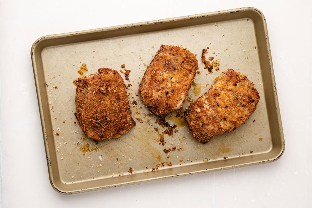 Three golden brown parmesan crusted pork chops on a baking sheet after cooking.