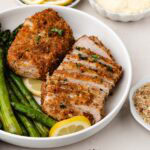 Sliced parmesan crusted pork chop on a plate next to asparagus and another pork chop.