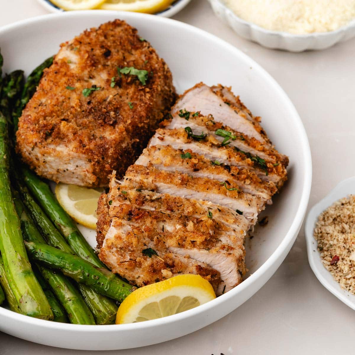 Sliced parmesan crusted pork chop on a plate next to asparagus and another pork chop.