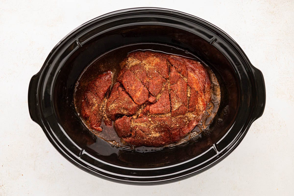 Slow cooker with the pork shoulder nestled in the braising liquid.