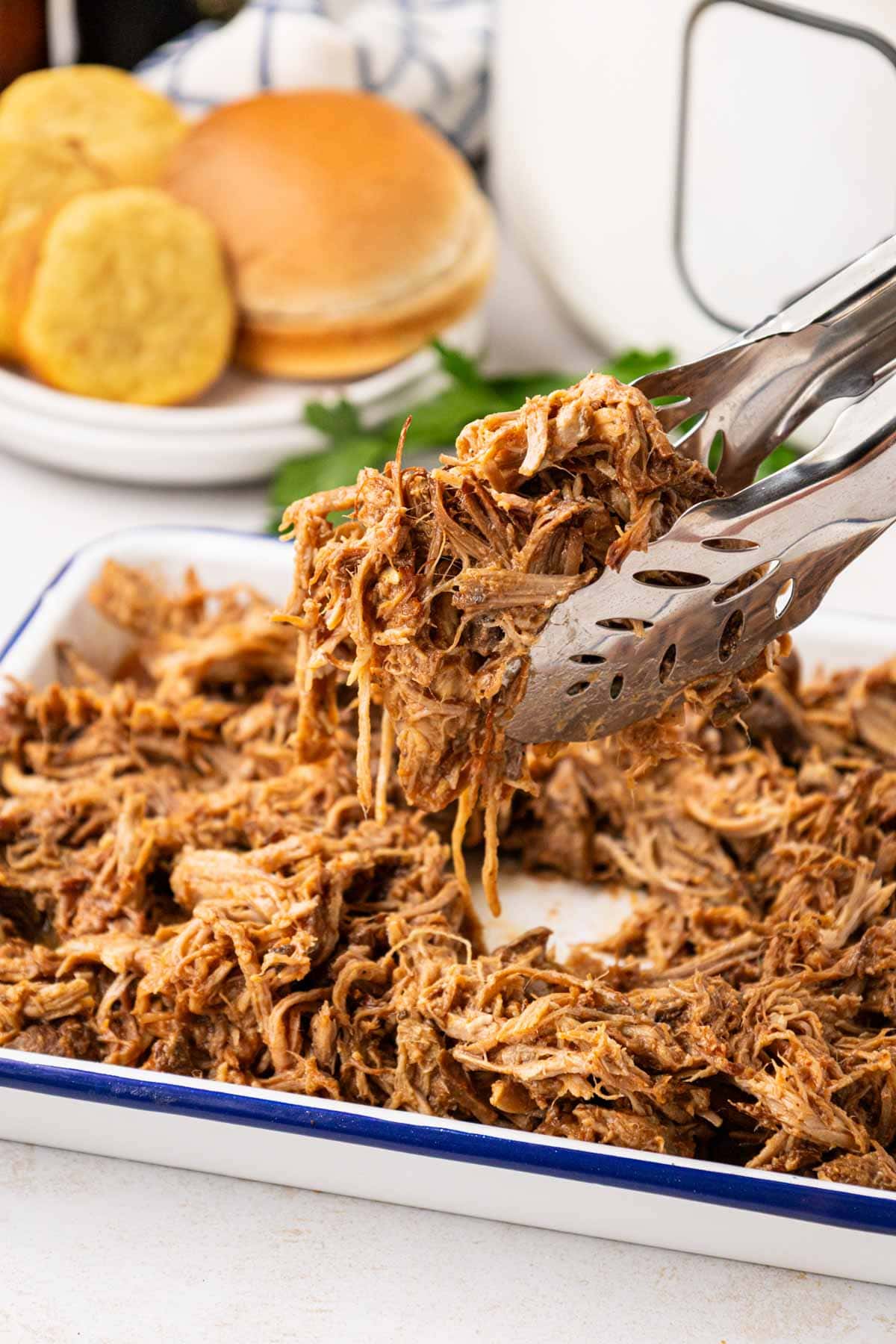 Tongs removing a serving of crock pot root beer pulled pork from serving tray.