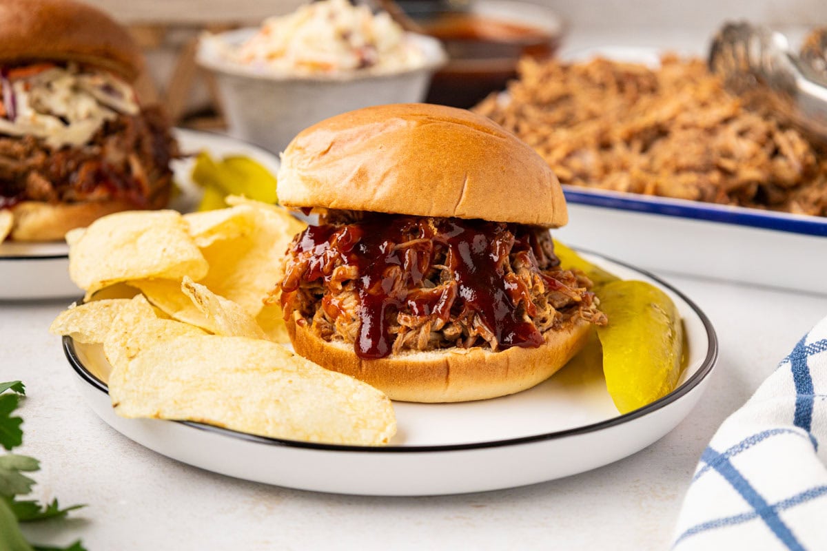 Root beer shredded pork sandwich on a bun served with pickles and chips.