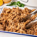 Tongs removing a serving of crock pot root beer pulled pork from serving tray.