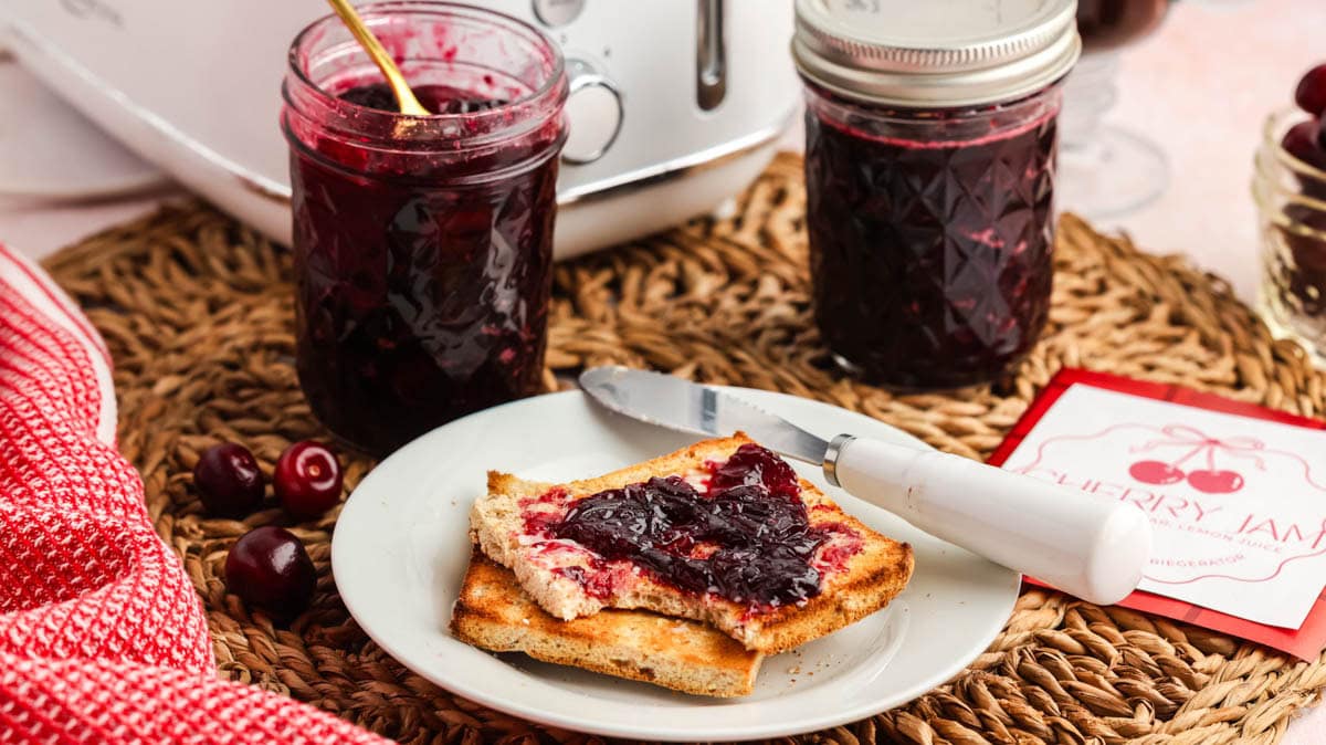 Cherry jam in jars next to some pieces of toast with jam on them.