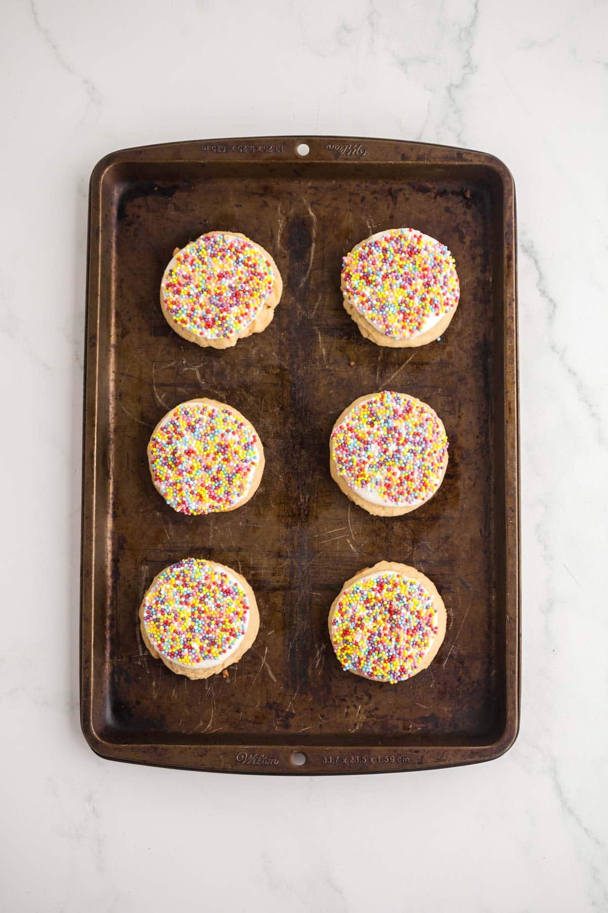 Icing and rainbow sprinkles on top of the sugar cookies on a sheet pan.