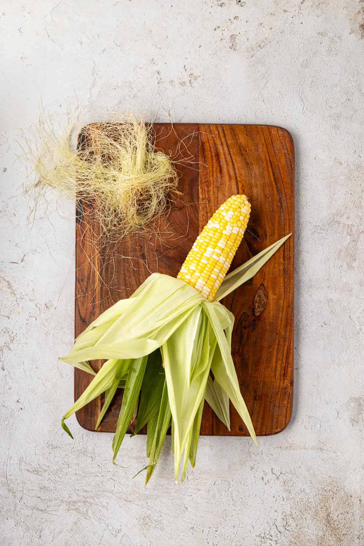Corn husk on a wooden board with husks pulled back and silk removed from the corn.