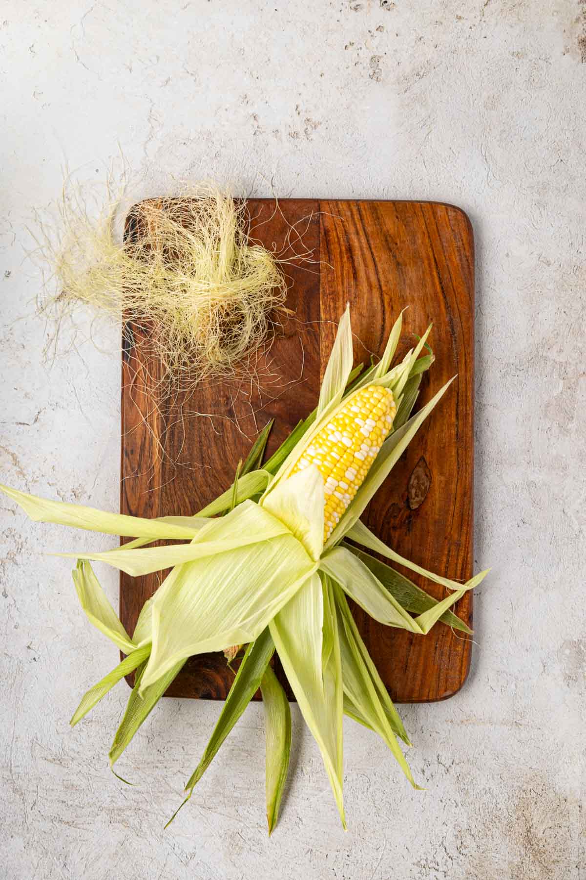 Corn husk on a wooden board with partial husk being pulled back up once silk removed.