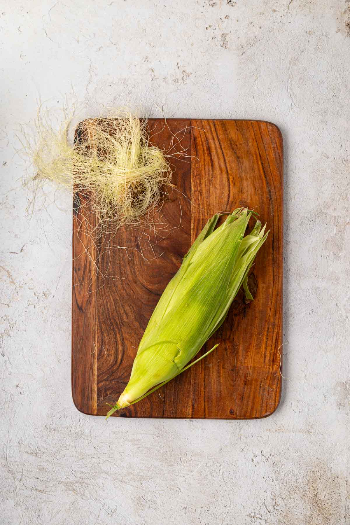 Corn husk on a wooden board with the entire husk being pulled back up over the corn once silk removed.