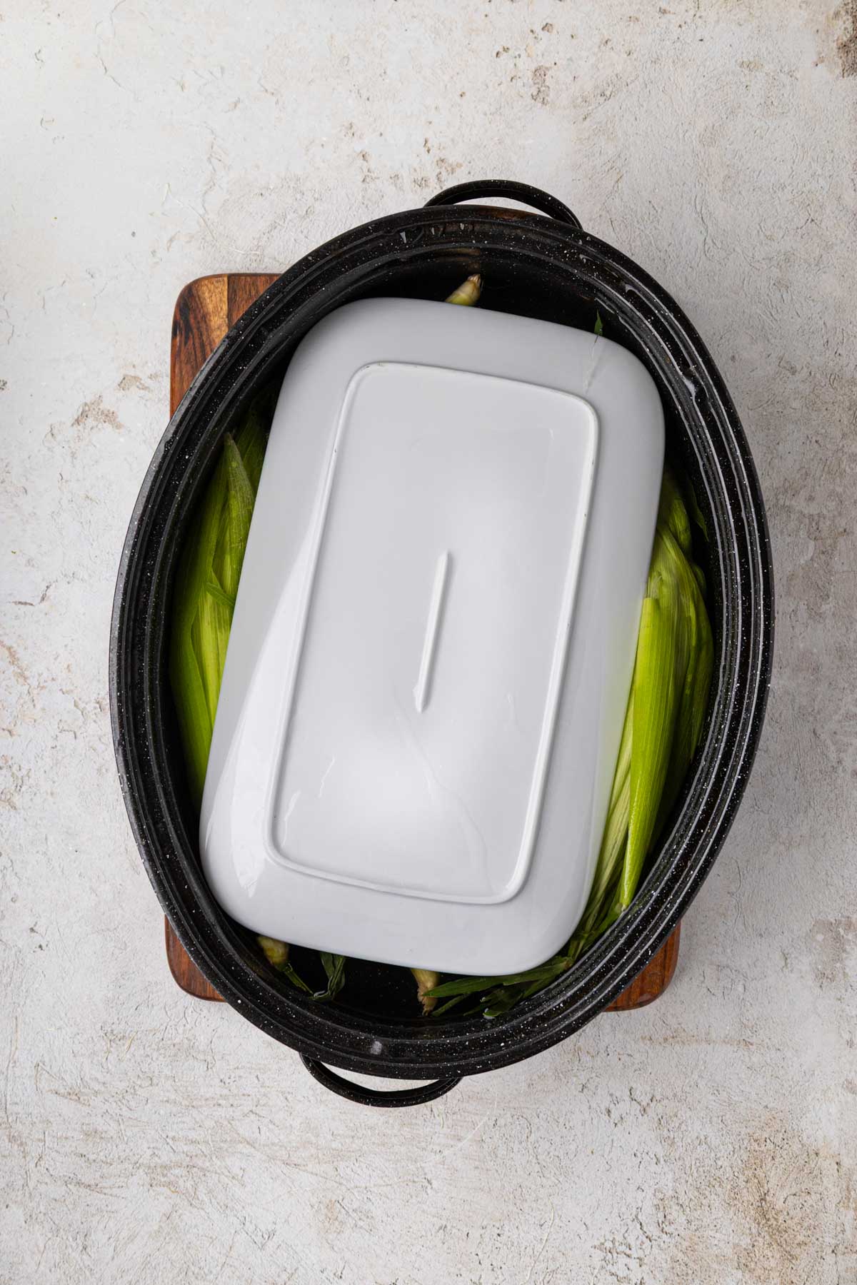Cleaned corn in the shuck in a pot of water with a plate holding them down in the water to soak.