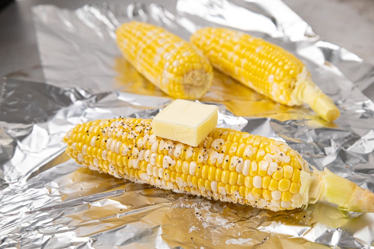 Corn cob sprinkled with salt & pepper an da pat of butter on top, sitting on a piece of foil.