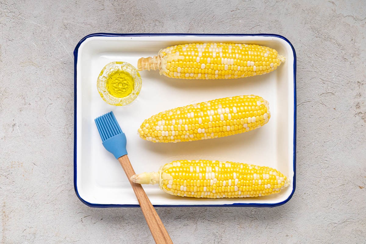 Shucked corn on a tray with a brush and oil.