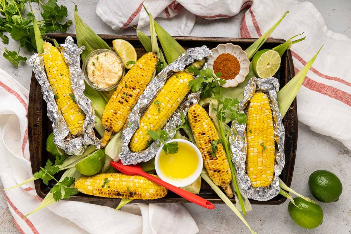 Grilled corn on the cob with some in the foil and some with charred corn from direct grilling on a platter with melted butter and seasonings.