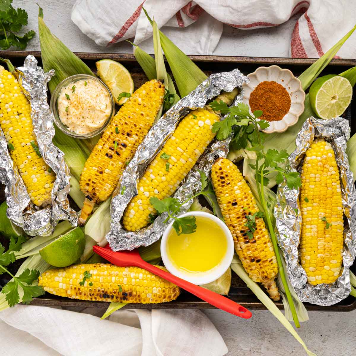 Grilled corn on the cob with some in the foil and some with charred corn from direct grilling on a platter with melted butter and seasonings.