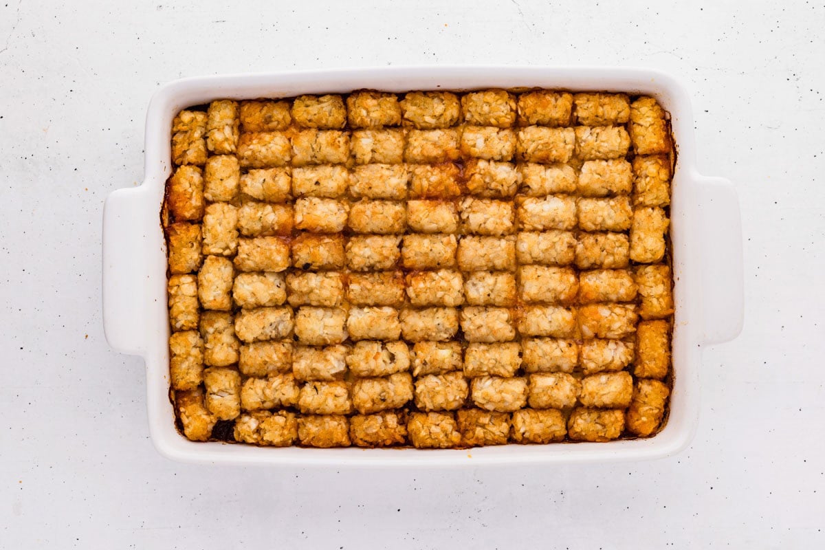Tater tots lined up on top of the sloppy joe and cheese mixture in a white baking dish.