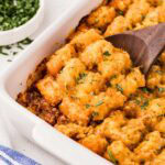Crispy tater tots on top of a sloppy joe mixture in a white baking dish with a wooden spoon.