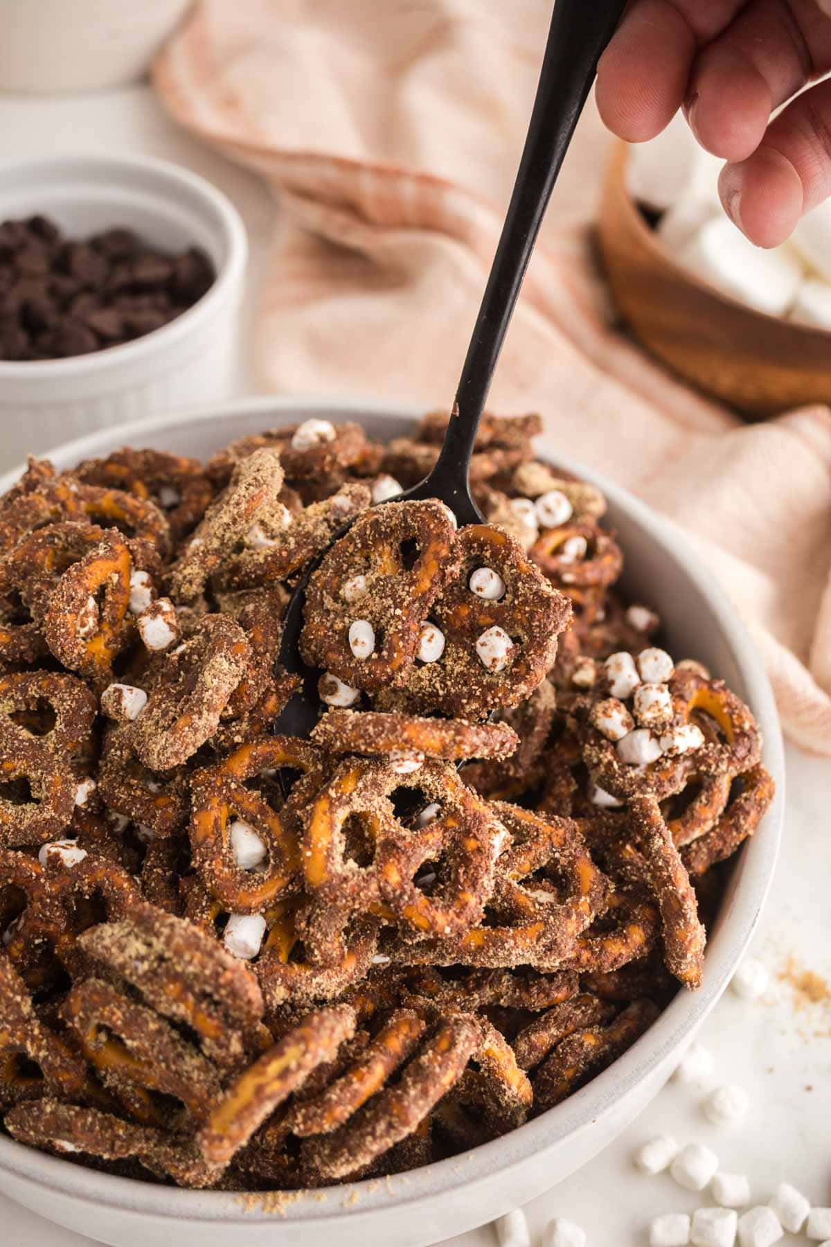 Hand holding a spoon, scooping up some s'mores pretzels from a bowl.